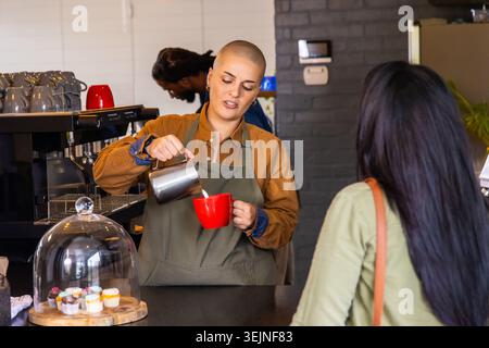 Female barista wearing olive apron pouring milk from metal pitcher into red mug at counter. Cafe, espresso, cupcakes, cloche, br Stock Photo