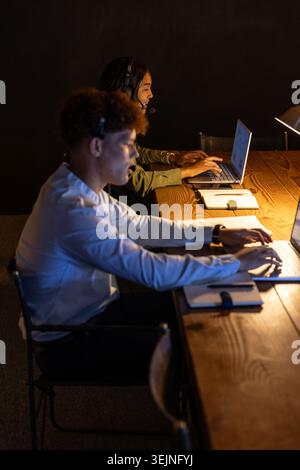 African American colleagues typing on laptops, speaking into headsets at call center desk with lamp. Professional, teamwork, col Stock Photo