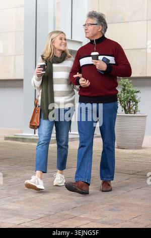 Senior couple walking arm-in-arm on paved plaza, holding coffee cups, bag, scarf, folded device. Urban, outdoor, elder, partners Stock Photo