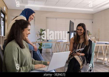 Diverse female coworkers collaborating on laptop and tablet in co-working room with blueprints. Collaboration, teamwork, minimal Stock Photo