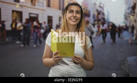 Woman holding yellow book, hands visible and smiling while reading on street; curiosity learning joyful. Stock Photo