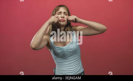 Young woman pinching her cheeks and pouting while making a sad face in a pink studio set; melancholy. Stock Photo