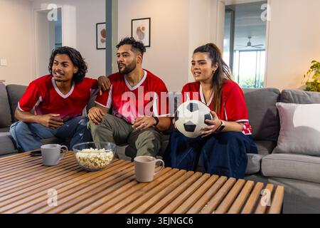 Asian friends in red jerseys watching match at home on sofa, woman holding soccer ball, popcorn. Watchparty, fans, football, uni Stock Photo