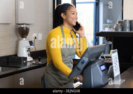 African American woman barista wearing gray apron speaking on phone behind counter by POS terminal. Cafe, coffee, daylight, modern, tidy, equipment, m Stock Photo