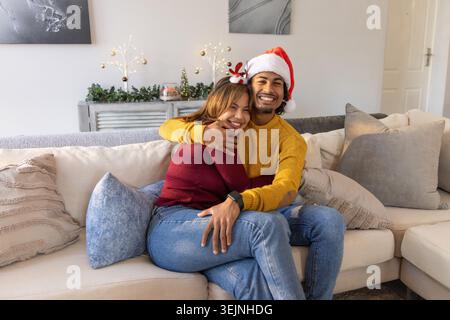 Couple hugging while sitting on sectional sofa at home wearing Santa hat and reindeer headband. Cozy, festive, holiday, romantic Stock Photo