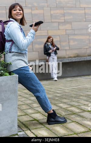 Two women leaning on stone planter in plaza, holding smartphones and purple backpack. Courtyard, urban, candid, casual, denim, c Stock Photo