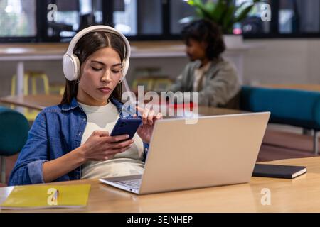 Diverse students studying in library, female with headphones using smartphone with laptop, notebook. Coworking, focus, workspace Stock Photo