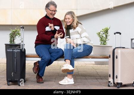 Senior couple sitting on bench at station, holding smartphone and coffee, black and beige suitcases. Retiree, duo, luggage, phon Stock Photo