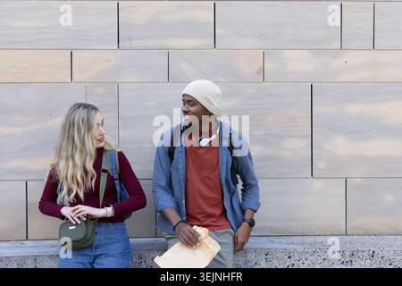 Diverse couple leaning on stone ledge, making eye contact, holding backpacks, envelope, headphones. Urban, plaza, campus, duo, casual, layered, bag Stock Photo