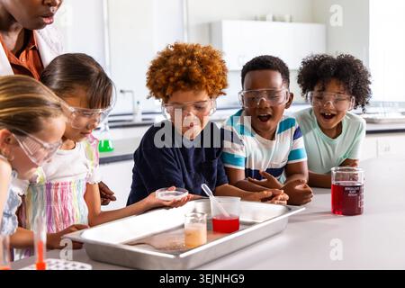 Diverse elementary kids wearing goggles crowding bench, stirring colored liquids near 600ML beaker. Students, classroom, science Stock Photo
