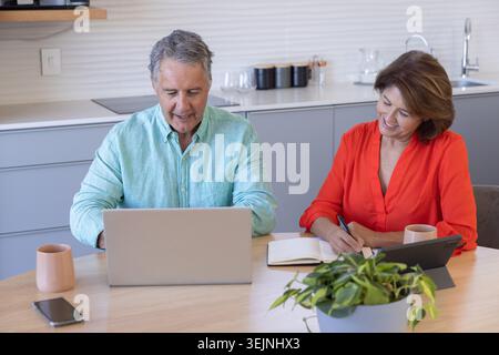Mature male and senior female couple sitting at kitchen table, using laptop and writing in notebook. Homeoffice, collaboration, productivity, remote, Stock Photo