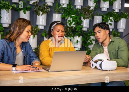 Diverse coworkers collaborating on silver laptop at modern co-working space with VR headset. Teamwork, collaboration, technology, design, innovation, Stock Photo