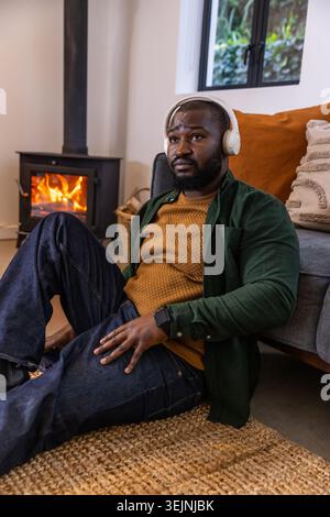 African American man sitting on jute rug at home wearing headphones, mustard sweater by wood stove. Cozy, livingroom, fireplace, Stock Photo