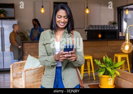 Indian woman standing near wooden cafe counter, smiling at smartphone, wearing olive-green shirt. Coffeehouse, phone, customer, Stock Photo