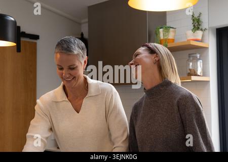 Senior female and woman smiling while cooking at kitchen counter with pendant lights, glass jar. Cozy, modern, home, countertop, Stock Photo