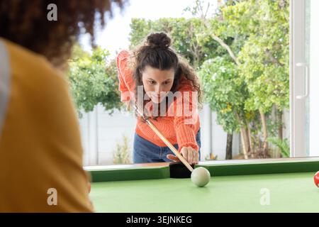 Female friends playing on pool table at home with woman in orange sweater aiming wooden cue. Billiards, backyard, daylight, casu Stock Photo