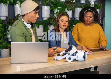 Diverse coworkers collaborating at counter in coworking space with VR headset, laptop, plant wall. Collaborative, design, office Stock Photo