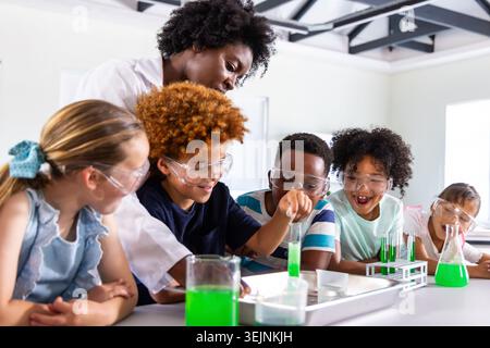 Female teacher guiding five children wearing goggles, pouring green liquid into test tubes in lab. Classroom, science, education Stock Photo