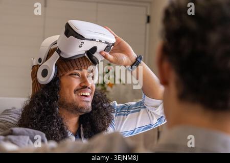 Diverse male friends sitting in living room, beanie wearing man holding white VR headset. Duo, cozy, gadget, wearable, casual, lounge, modern Stock Photo