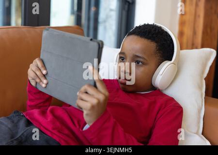 African American child reclining at home on sofa using gray tablet wearing red shirt, white headset. Couch, headphones, screen, Stock Photo