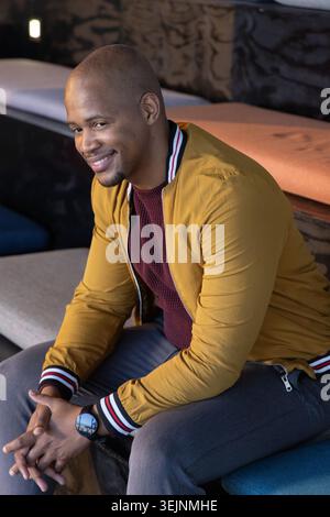 African American man sitting on bench in lounge wearing mustard bomber jacket and smartwatch. Interior, cafe, cozy, warm, relaxe Stock Photo