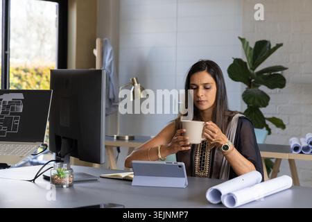 Indian woman wearing kurta sitting at desk in studio holding mug reviewing tablet plan monitor. Architecture, design, office, wo Stock Photo