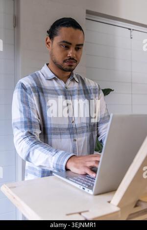 Asian man typing on silver laptop, standing at desk wearing plaid shirt in office with plant. Adjustable, easel, workstation, mi Stock Photo