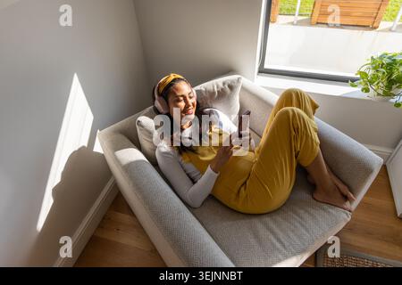 Asian woman reclining on gray chair at window in yellow jumpsuit with headset holding phone smiling. Sofa, light, sunlit, minima Stock Photo
