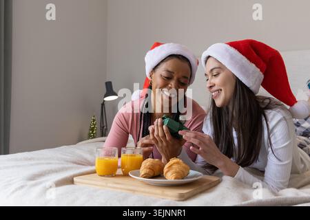 Diverse female friends reclining on bed wearing Santa hats, smiling and examining gift with tray. Cozy, festive, morning, compan Stock Photo