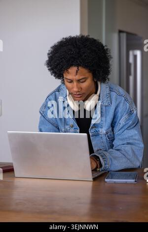 African American woman typing on silver laptop at kitchen table wearing denim jacket headphones. Workspace, remotework, homeoffi Stock Photo