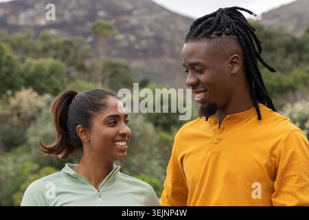 Diverse couple smiling and facing each other while standing on trail in hills wearing activewear. Nature, outdoor, overcast, hiking, companionship, pa Stock Photo