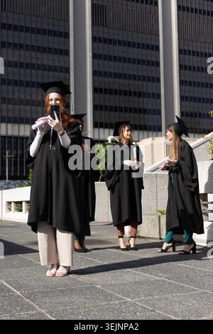 Diverse female graduates taking phone selfie, holding diplomas in gowns and caps on urban plaza. Diploma, mortarboard, celebrati Stock Photo