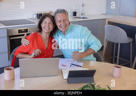Senior woman in red blouse and man in blue shirt reviewing laptop charts at kitchen table. Couple, dining, tablet, graphs, colla Stock Photo