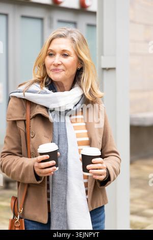 Middle-aged woman standing on city sidewalk holding two takeaway coffee cups wearing tan coat scarf. Urban, storefront, glass, entrance, overcast, cas Stock Photo