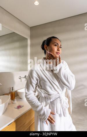 Asian woman wearing bathrobe, standing in bathroom beside vanity and mirror, checking skincare. Minimalist, serene, washroom, ro Stock Photo