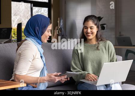 Diverse female colleagues discussing on gray sofa in workspace with tablet, laptop, hijab, sweater. Collaboration, mentorship, d Stock Photo