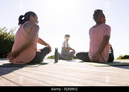 Senior woman leading women in activewear doing seated twists on deck with yoga mats, water bottle. Trio, platform, outdoor, sunl Stock Photo