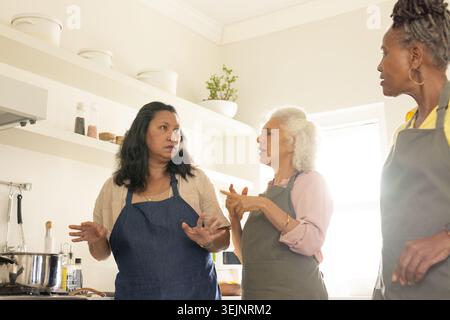 Senior and mature female friends wearing aprons cooking together in home kitchen with pot and tools. Trio, women, sunlit, stovetop, utensils, cozy, ca Stock Photo
