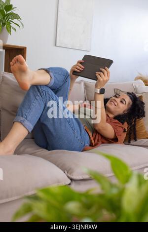 Woman reclining on gray sofa holding tablet overhead, reading during home break with smartwatch. Device, plants, casual, relaxed Stock Photo