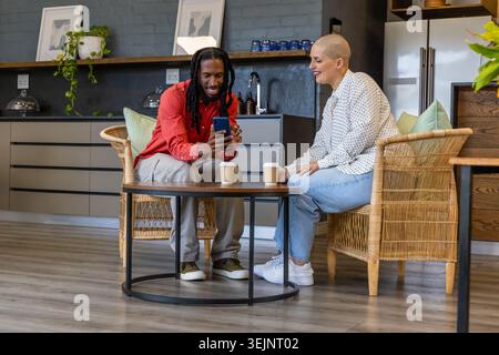 Diverse coworkers sitting in kitchen lounge sharing smartphone and takeaway cups on wooden table. Colleagues, conversation, phon Stock Photo