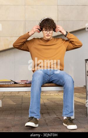 Teenage male adjusting over-ear headphones while sitting on bench in plaza with notebooks and watch. Youth, student, outdoor, urban, courtyard, daylig Stock Photo
