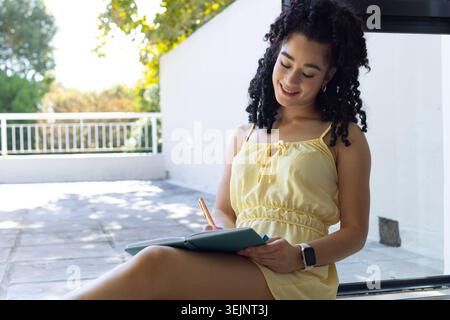 Woman 20s writing in notebook with pen on patio wearing sundress, sunlight inspiring, copy space. Terrace, journal, teal, sunshi Stock Photo