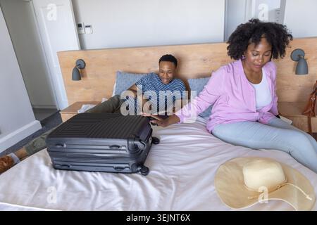 African American couple sitting on bed, woman reaching toward black suitcase with straw hat. Luggage, hotel, duo, travel, getawa Stock Photo