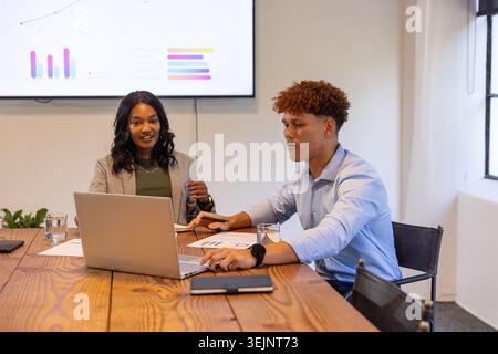 Diverse coworkers analyzing data on laptop with charts and mounted display at office boardroom. Collaboration, teamwork, analytics, corporate, modern, Stock Photo