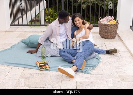 Diverse couple sitting on blue picnic blanket on stone patio, woman in white camisole, sipping wine. Outdoor, romance, terrace, Stock Photo