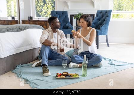 Diverse couple sitting on light-blue blanket in bright bedroom, sharing smartphone and sipping wine. Cozy, homey, interior, sunl Stock Photo