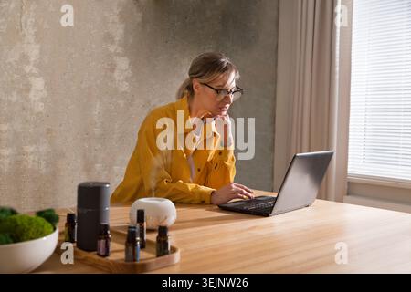 Woman working on laptop from home near essential oil aroma diffuser humidifier diffusing water articles in the air. Aroma therap Stock Photo