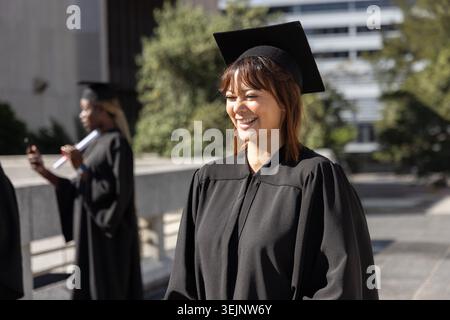 Female graduate smiling and posing on campus walkway wearing gown holding cap and diploma. Graduation, celebration, sunny, outdoor, university, classm Stock Photo