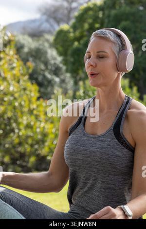 Middle-aged woman sitting meditating in park wearing sportswear, pink headphones and smartwatch. Outdoor, nature, grass, foliage Stock Photo