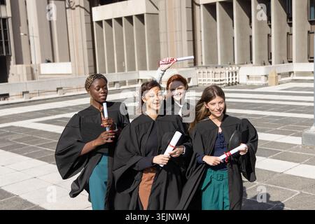 Diverse female graduates walking across campus plaza in gowns, holding diplomas and caps, smiling. Commencement, ceremony, morta Stock Photo